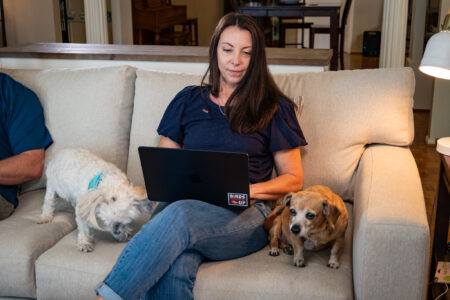 A woman holds a laptop in her lap while sitting on a beige couch between two dogs.
