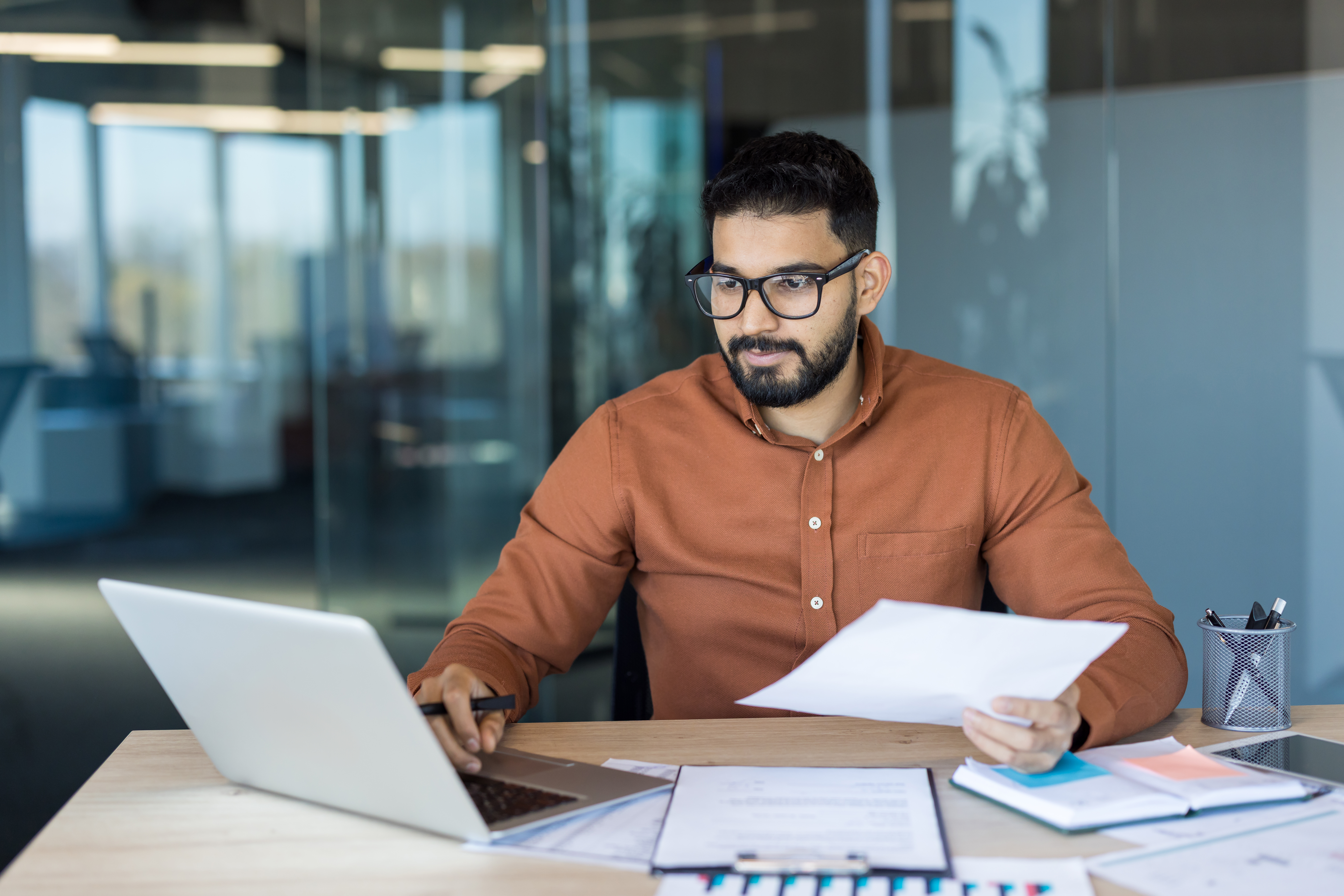 Man with glasses and an orange shirt sitting in an office working on a laptop.