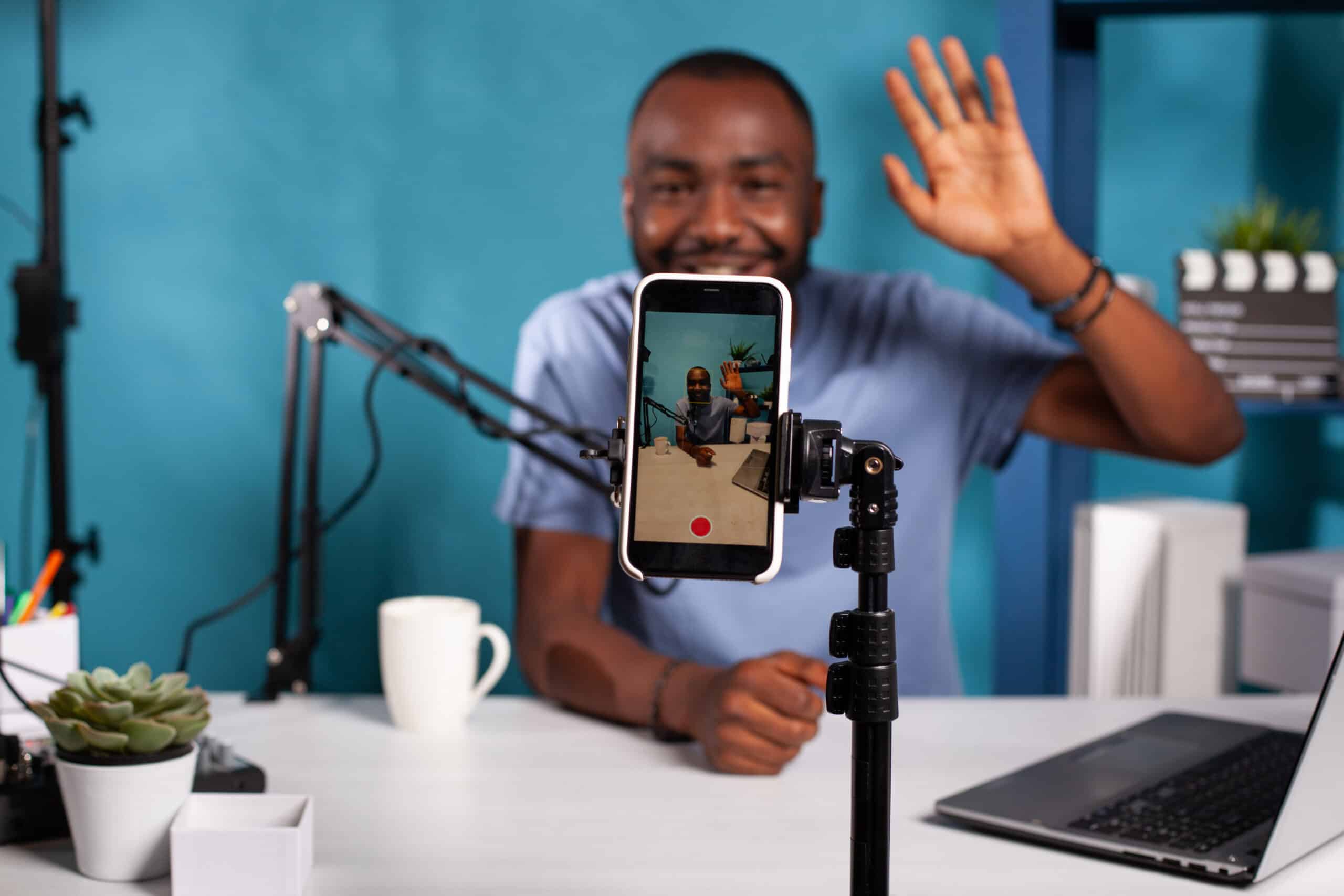 Closeup of live vlog setup with smartphone on stand filming vlogger waving hello sitting at desk with professional microphone.