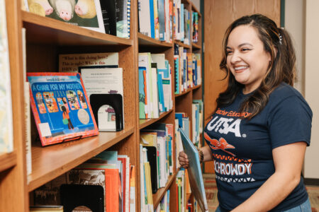A teacher with a blue UTSA t-shirt on putting a children's book back on the bookshelf.