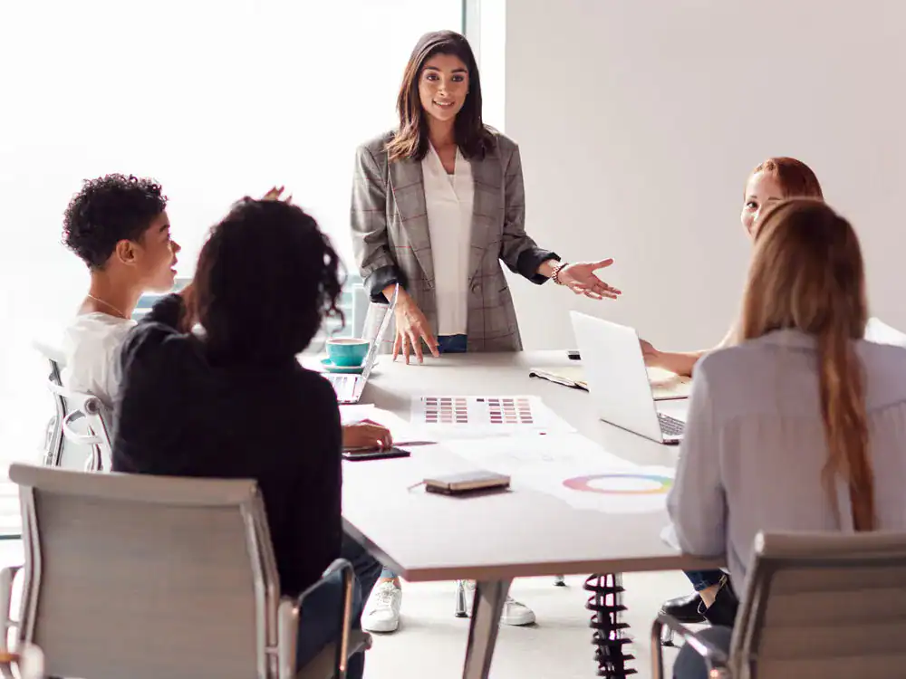 Woman in a white shirt and beige blazer speaks to a table of students.