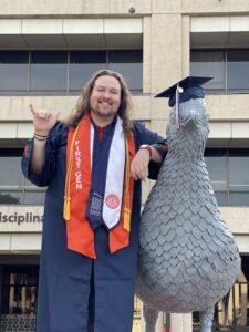 Man wearing graduation regalia standing next to roadrunner statue on UT San Antonio's campus.