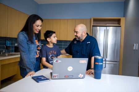Three people in a kitchen setting engage in conversation around a laptop adorned with stickers with a dark blue tumbler and notebook nearby.