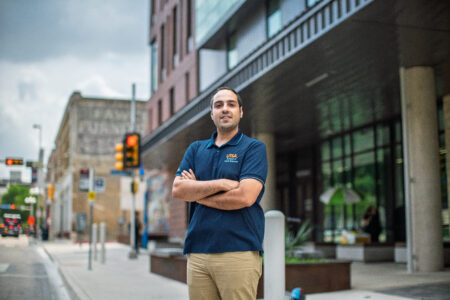 Man wearing a blue polo and khaki pants standing on the edge of a downtown street with his arms crossed.