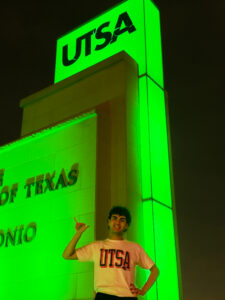 UT San Antonio Online graduate Anthony Montes Nieves stands in front of the UTSA front entrance sign holding up the "Roadrunner" hand sign.