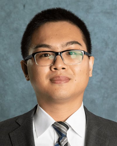 Man wearing glasses and a suit in front of a gray background.