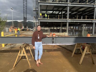 Woman wearing jeans and brown sweater stands in front of a large steel beam at a construction site.
