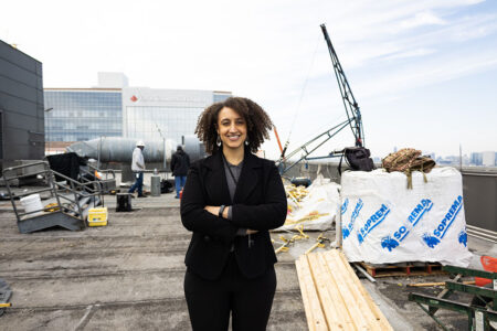 Woman wearing a black suit stands with her arms crossed on the roof of a building with construction equipment around her.
