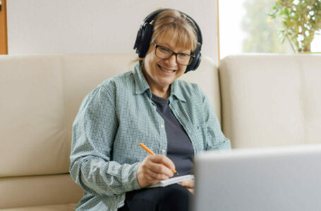 Modern middle aged blond woman in headphones take online course or training on computer at home. 